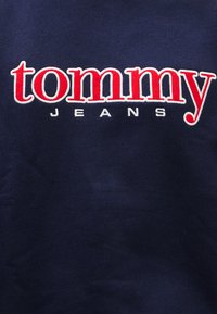 Tmavomodrá látka s rebrovanou textúrou, uprostred výrazne vyšitá červeným logom „tommy“ a bielym nápisom „JEANS“.