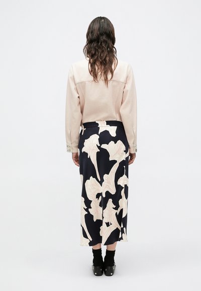 Μπεζ τοπ με μακριά μανίκια και εμφανείς ραφές, συνδυασμένο με μια ναυτική φούστα με ροζ λουλουδάτο σχέδιο και μαύρα παπούτσια με μεταλλικές λεπτομέρειες.