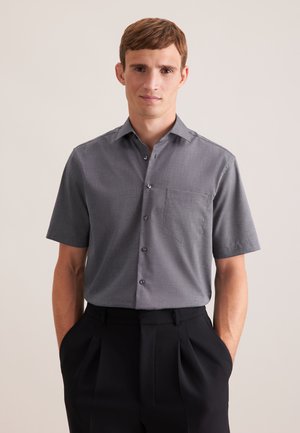 Mand iført grå kortærmet skjorte med brystlomme og sorte plisserede bukser, hænder i lommerne, neutral baggrund.