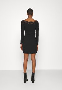 Svart långärmad klänning med spetstydlighet längs axlarna, figurnära silhuett och rynkad sida, kombinerad med svarta ankelstövlar.