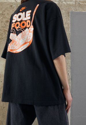 Person iført sort skjorte med orange Nike-logo og teksten "SOLE FOOD", med sneakers- og ske-grafik på bagsiden.