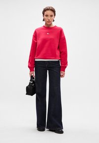 Sweatshirt rouge à col rond, associé à un pantalon évasé en denim foncé. Sac à main noir avec poignée, texture lisse et détails minimalistes.