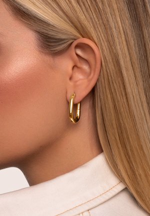 Krupni plan ženinog uha s glatkom kožom i ravnom plavom kosom koja nosi sjajnu zlatnu naušnicu u obliku obruča i kremastu košulju s ovratnikom.