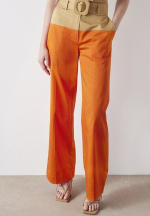 Henkilö, jolla on päällään korkeavyötäröiset, leveälahkeiset oranssit housut, joissa on beige vyö, jossa suuri pyöreä solki, sekä remmikkäät neliökärkiset sandaalit.