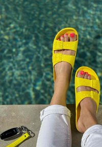 Sandales à paillettes jaunes avec deux boucles réglables, posées sur une surface en pierre près d'une piscine. Pantalon blanc retroussé et ongles de pieds roses visibles.