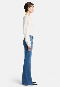 Top bianco a maniche lunghe con scollo a V, abbinato a jeans blu a vita alta e svasati. Scarpe nere con punta a forma di freccia, modella in posizione di profilo.