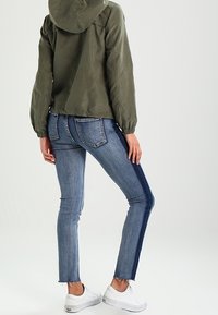 Chubasquero verde oliva con capucha y puños elásticos, combinado con jeans ajustados azules con dobladillo deshilachado y una raya oscura a los lados. Zapatillas blancas.