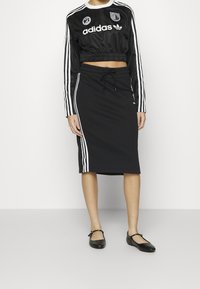 Kvinna klädd i en svart Adidas kortärmad sweatshirt med vita ränder och en matchande svart kjol med vita ränder samt svarta Mary Jane-skor.