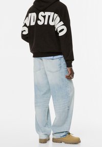 Personne portant un sweat à capuche noir avec un grand texte blanc au dos, un jean ample bleu clair, des bottes beige, ainsi que des bagues et un bracelet argentés.