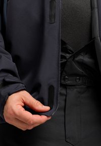 Giacca impermeabile nera con zip frontale, caratterizzata da una texture liscia, dettagli in velcro e un colletto aderente. Mano che tiene il tessuto.