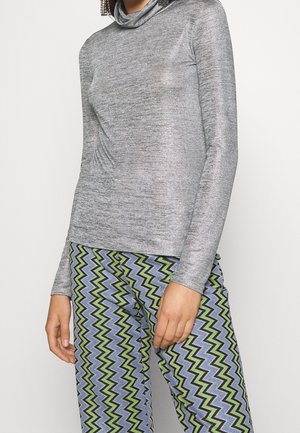 Kobieta w długim rękawie srebrnym golfie i wysokich, zygzakowatych spodniach w zielonym, niebieskim, czarnym i białym kolorze.