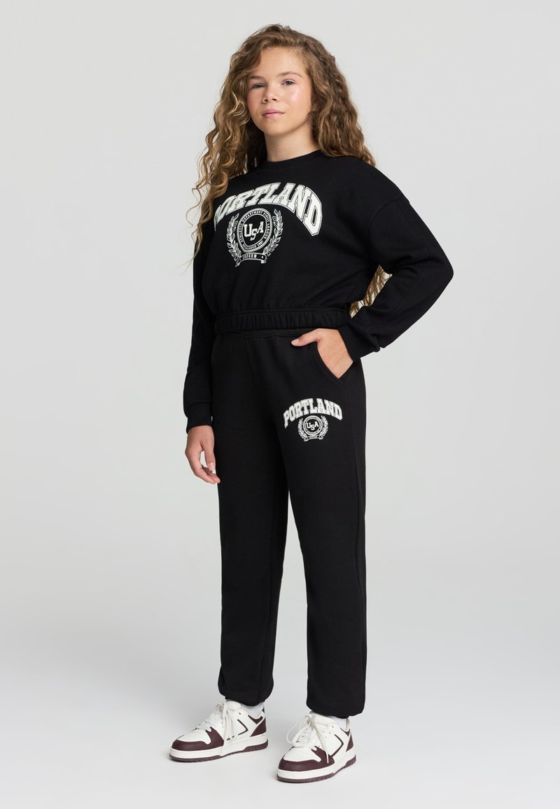 Czarny krótki sweatshirt z nadrukiem "PORTLAND" i logo w białym kolorze; zestawiony z czarnymi spodniami dresowymi z tym samym nadrukiem i białymi akcentami.