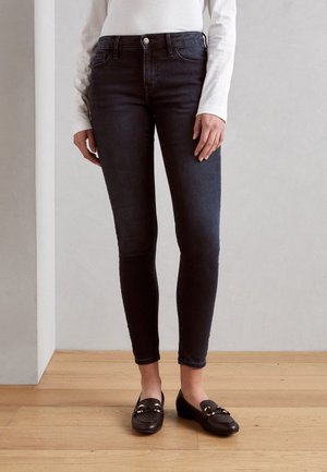 Jeans skinny de mezclilla oscura con cintura alta, con cierre de botón en la parte delantera y bolsillos. Combinados con mocasines negros adornados con detalles dorados.