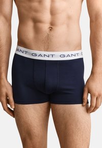 Torse masculin portant un boxer bleu marine avec une ceinture blanche portant la marque "GANT" en lettres bleu marine.