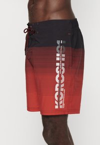 Costumi da bagno da uomo in gradiente da rosso a nero, realizzati in tessuto leggero, con strisce orizzontali e grande dettaglio del logo bianco sul lato.