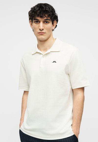 Νέος άνδρας με κυματιστά σκούρα μαλλιά, φορώντας λευκό πόλο μπλουζάκι με μικρό μαύρο λογότυπο, στέκεται με τα χέρια στις τσέπες σε απλό φόντο.