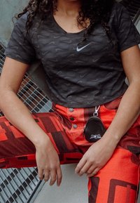 Svart Nike t-shirt med vågmönster, kombinerad med jeans med röd och svart grafisk tryck. Inkluderar en svart accessoarväska med dragkedja fäst vid byxorna.