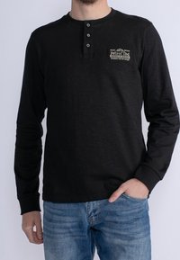 Mustapaita pitkällä hihalla, jossa on pyöreä kaula-aukko, kolmesta napista koostuva nappilista ja edessä beige logo. Pehmeä rakenne, istuva muotoilu ja rento tyyli.