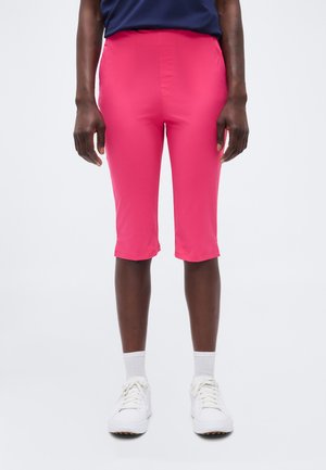 Persoană purtând pantaloni scurți roz aprins până la genunchi, șosete albe, adidași albi și un tricou albastru închis, stând în picioare pe un fundal simplu.