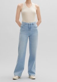 Persona que lleva una camiseta de punto sin mangas color beige, jeans de pierna ancha de cintura alta en azul claro y zapatillas blancas, con las manos detrás de la espalda.