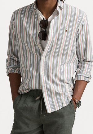 Mann trägt ein gestreiftes Hemd mit hochgekrempelten Ärmeln, dunkle Sonnenbrille hängt am Hemd, dunkle Hose und eine Armbanduhr.