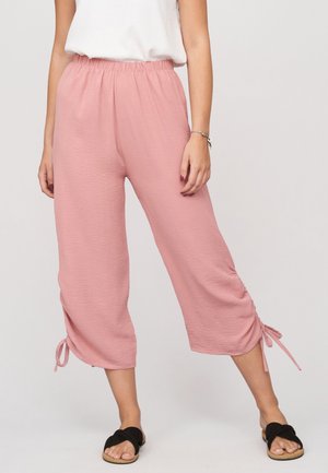 Mujer con pantalones cortos holgados rosas con cordones en el dobladillo, una camiseta sin mangas blanca, sandalias negras y una pulsera en la muñeca.