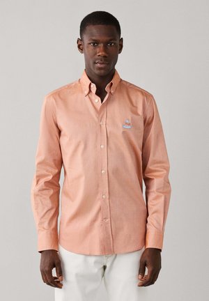 Hombre con camisa de manga larga color melocotón con un pequeño logo bordado en el pecho y pantalones blancos, de pie frente a un fondo liso.