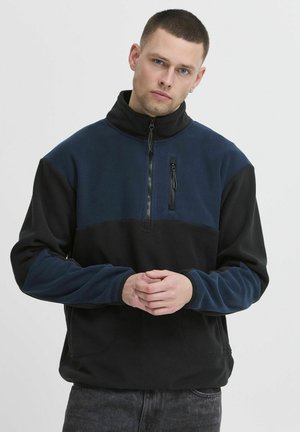 Sudadera de forro polar en azul oscuro y negro, con diseño de media cremallera y un bolsillo delantero con cremallera, además de dos bolsillos laterales. Textura suave y corte relajado.