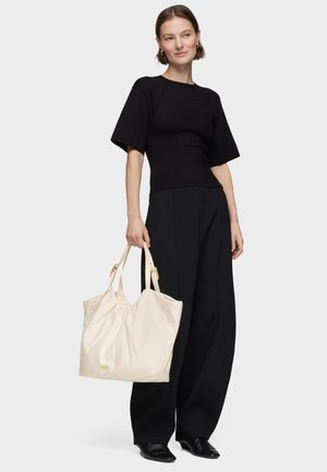 Mujer vestida con top negro y pantalones de pierna ancha, sosteniendo un gran bolso beige con doble asa, posando contra un fondo liso.