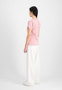 Light pink t-shirt with short sleeves, fitted design, and a logo tag on the left sleeve, paired with white, loose-fitting pants.