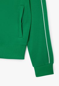 Grøn sweatshirt med ribbede kanter og ærmer, med en lynlåslomme og hvid piping langs ærmerne. Glat tekstur.