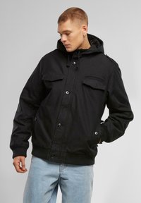 Black bomber jacket with a hood, featuring two chest pockets, snap closures, ribbed cuffs, and a soft texture. Worn with light blue jeans.