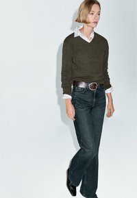 Maglione in maglia verde scuro, camicia bianca con colletto sotto, abbinato a jeans a zampa d'elefante in denim scuro e cintura in pelle marrone. Insieme smart-casual.