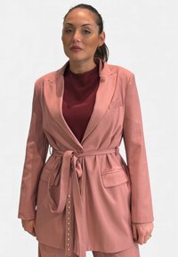 Blazer rosa con cintura, dal design su misura, caratterizzato da un unico bottone frontale, due tasche e bottoni decorativi lungo la cucitura laterale.