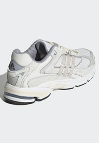 Fehér sportcipő hálós és velúr anyagból, három szürke csíkkal, párnázott talppal, párnázott szegéllyel és texturált részletekkel a hátsó részen.