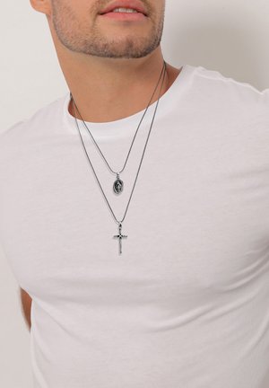 Mand i hvid skjorte iført to sølvhalskæder, den ene med en religiøs medaljon og den anden med et korsvedhæng.