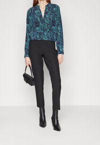 Blouse imprimée en bleu sarcelle foncé et marine avec un décolleté en V, manches longues ; associée à un pantalon noir ajusté et des bottes noires jusqu'aux genoux. Petit sac à main noir.