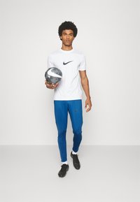 Weißes Nike-T-Shirt mit navy Logo, blaue Nike-Hose mit strukturiertem Design, hält einen schwarz-silbernen Fußball, trägt schwarze Fußballschuhe.