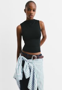 Schwarzes ärmelloses Oberteil mit hohem Kragen, kombiniert mit einem braunen Gürtel über dunklen Jeans und einem geknoteten grün-weiß gestreiften Hemd um die Taille.