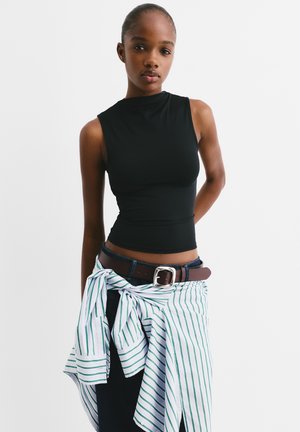 Schwarzes ärmelloses Oberteil mit hohem Kragen, kombiniert mit einem braunen Gürtel über dunklen Jeans und einem geknoteten grün-weiß gestreiften Hemd um die Taille.