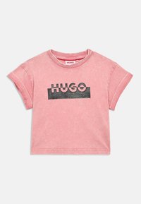 Rozā īsām piedurknēm T-kreklā no mīksta auduma, ar melnu "HUGO" logotipu uz krūšu daļas, ar izbalējušu, nedaudz teksturētu izskatu.