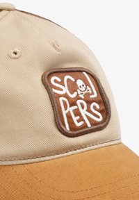 Gorra marrón con un parche beige que presenta texto blanco y un diseño de calavera. La tela tiene un acabado texturizado, con un ala curvada y detalles de costura.