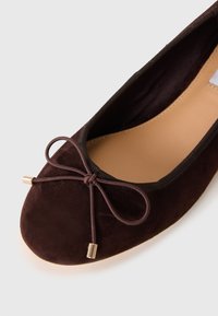 Dark brown suede ballet flat with a tied bow and gold metal tips on the toe, beige insole, and black trim around the opening.