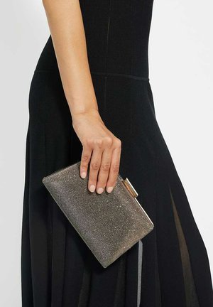 Pochette métallique avec une surface texturée, dotée d'un fermoir. La couleur est un argent-or brillant, tenue dans une main contre une robe noire.