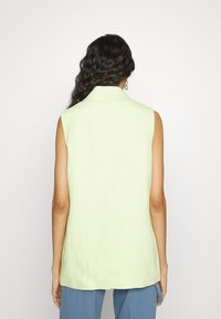 Jasnozielona, bezrękawnikowa marynarka z usztywnionym kołnierzem i prostym krojem, charakteryzująca się gładką fakturą oraz rozcięciem z tyłu dla swobody ruchów.