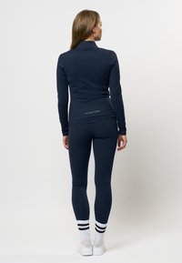 Vrouw met lang haar staat met haar gezicht naar weg, gekleed in een marineblauwe sportjack en leggings, witte sokken met zwarte strepen en witte sneakers.