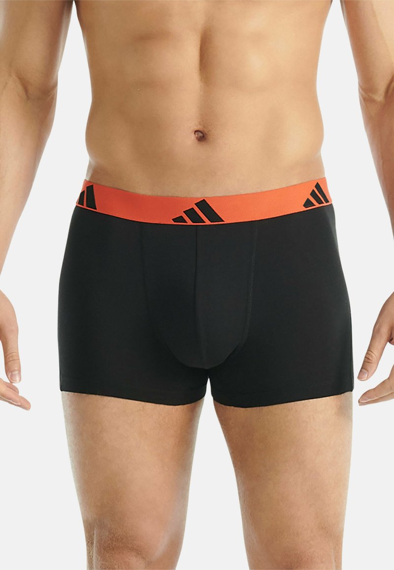 Torso et parties supérieures des jambes d'un homme portant un boxer noir avec une ceinture orange ornée de logos triangulaires noirs.
