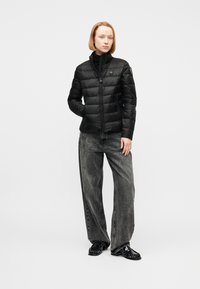 Femme aux cheveux courts et roux portant une doudoune noire, un jean gris à jambes larges et des mocassins noirs, debout avec une main dans la poche.