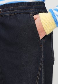 Temno jeans hlače imajo bočne žepe, kontrastne oranžne šive in elastičen pas. Tkanina ima gladko teksturo in rahlo sijoč videz.