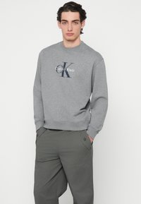 Šedá mikina s okrúhlym výstrihom, na ktorej je veľké tmavomodré logo Calvin Klein. Doplnok tvoria voľné olivovo zelené nohavice s vreckami.